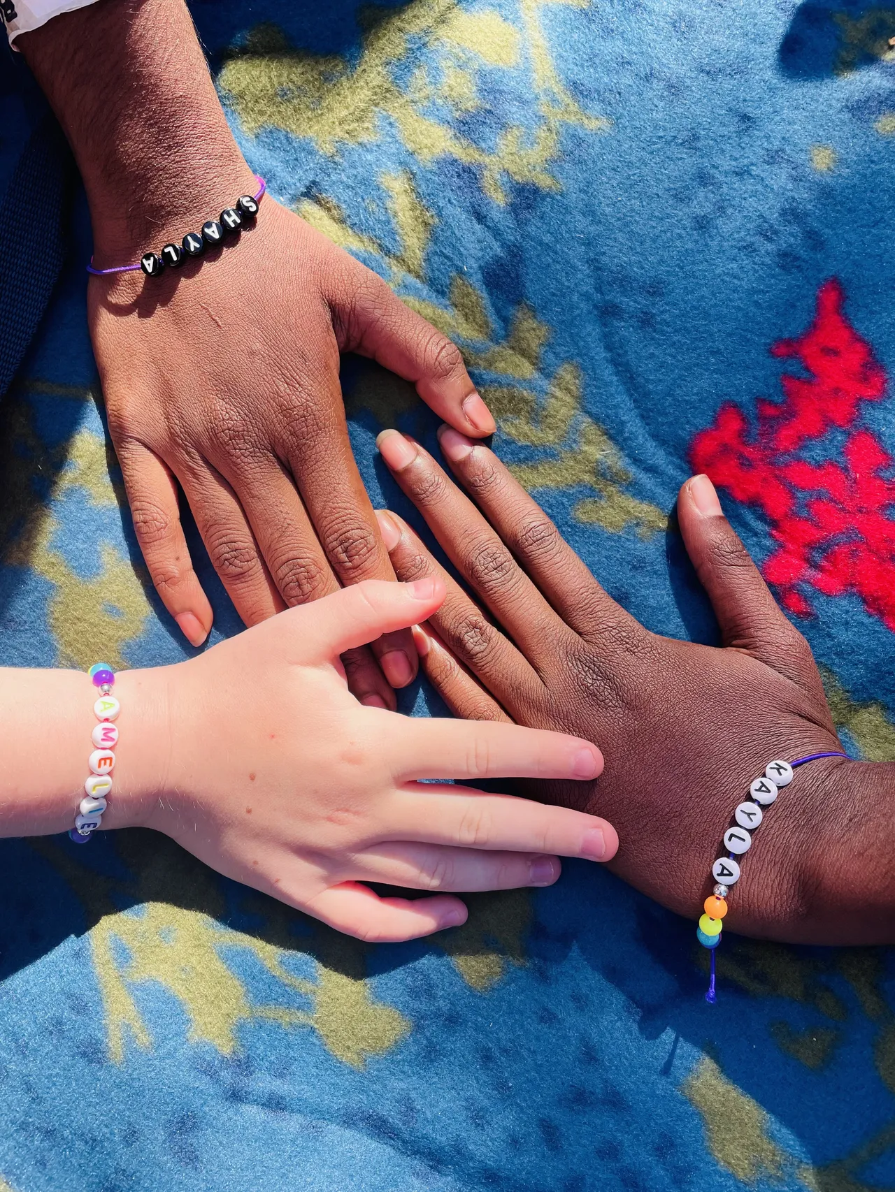 A close-up photo of three hands with different skin tones, gently touching over a vibrant mat. Each wrist adorned with a friendship bracelet, symbolising unity and friendship across diverse backgrounds.