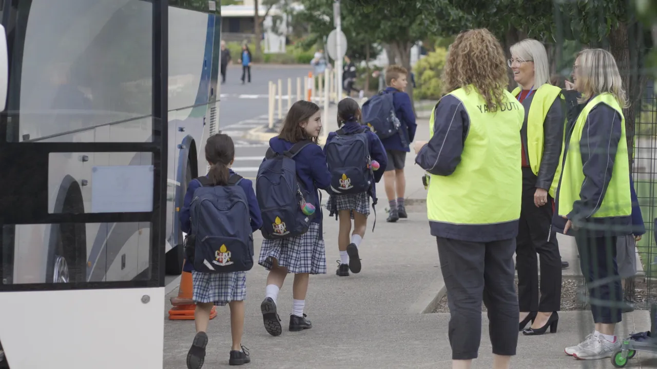 Lower primary school aged girl smiling to staff on yard duty as he gets off the bus in the morning at Canowindra Campus.