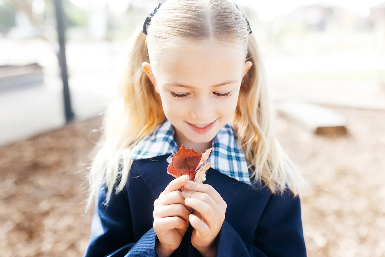 Lower primary school aged student from Overnewton's Junior School smiling as she closely inspects an autumnal leaf with both hands.