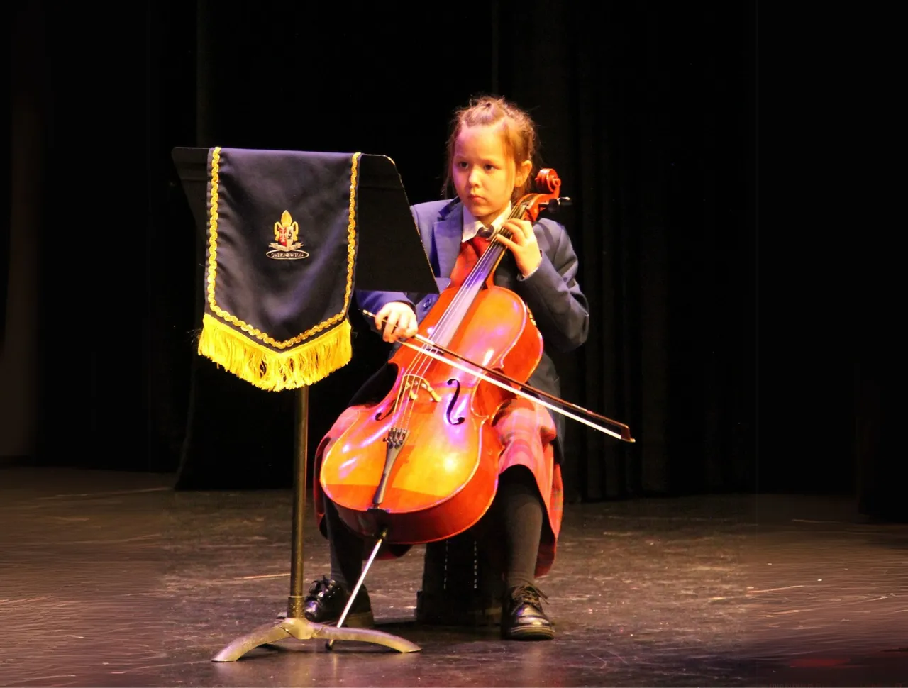 Lower primary school aged student playing the cello with deep concentration on stage, as part of an official musical showcase at Overnewton’s annual Festival of Music.