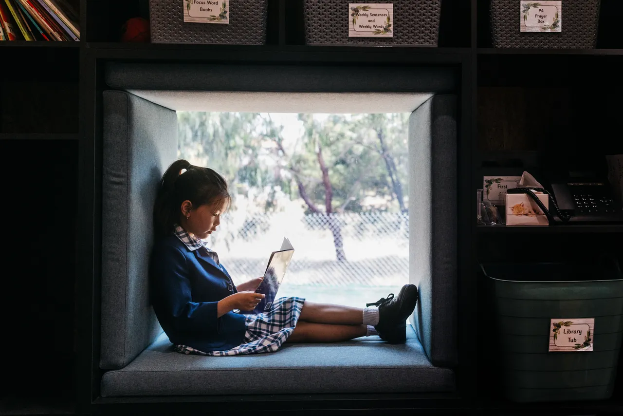 Lower primary school aged student tucked away in a cosy window reading nook at Canowindra Campus, reading a book with trees and natural greenery visible through the window.