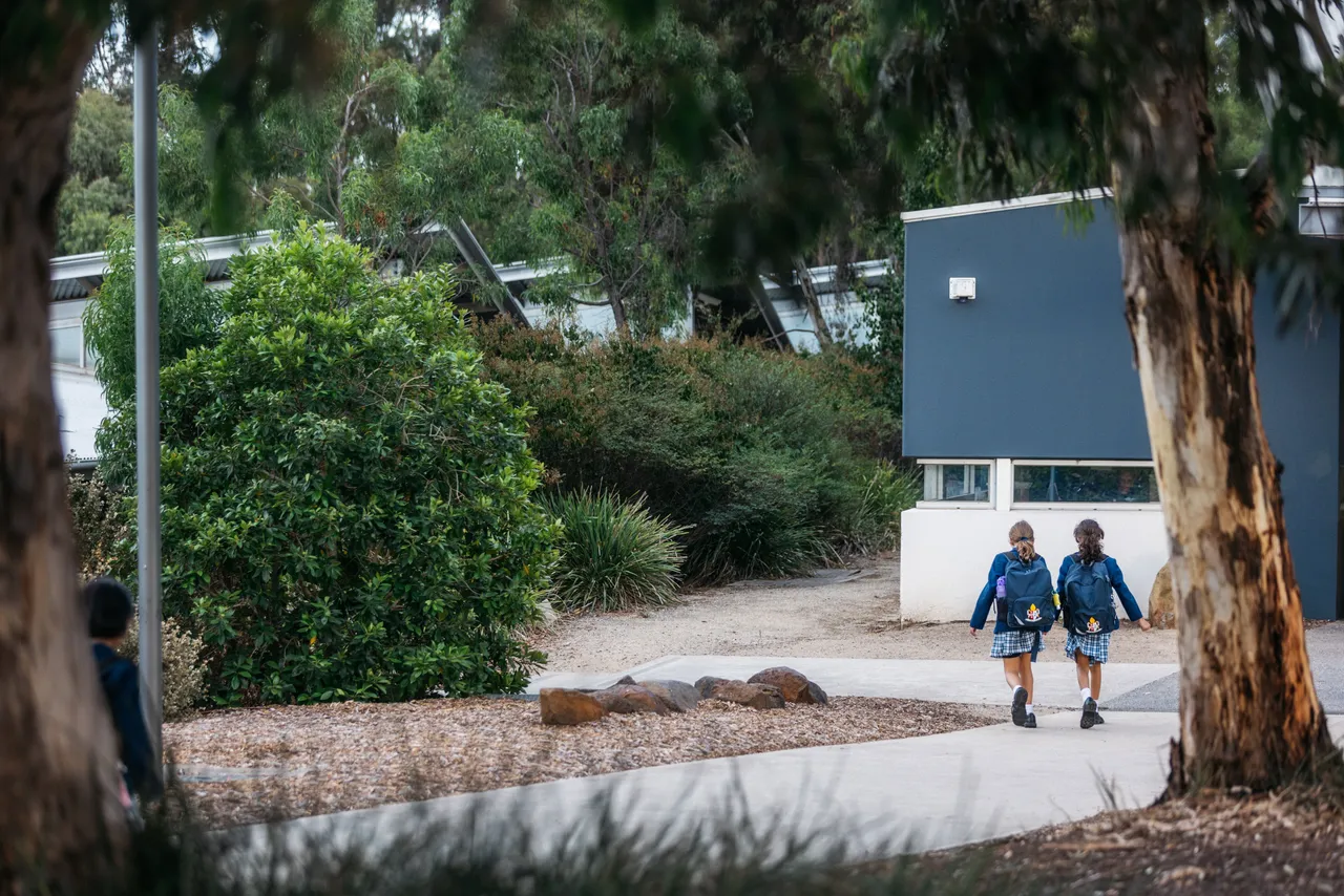 Two lower primary school aged students walking to class at Canowindra Campus, surrounded by trees and natural landscape, walking past the Junior School gymnasium.