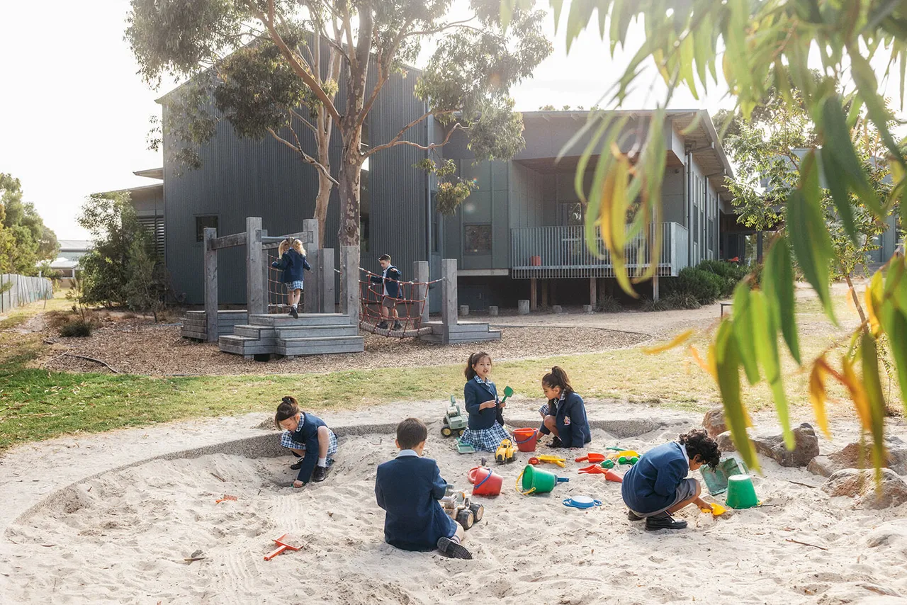 Seven primary school aged children playing in the sandpit and on the playground at Canowindra Campus, with trees, natural surroundings, and the Prep to Year 2 building in the background.