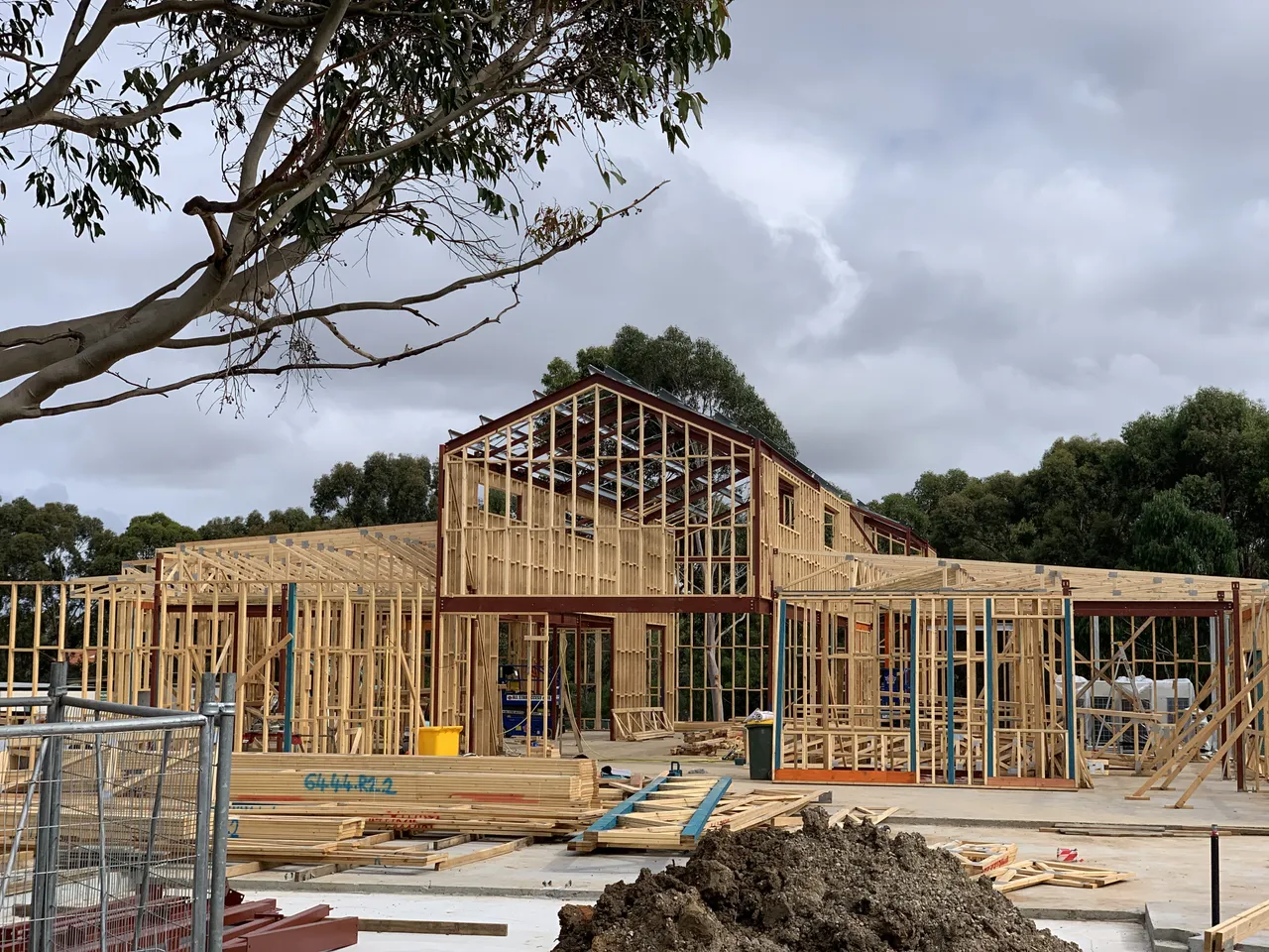 A large timber-framed building is under construction on a concrete slab, with scaffolding, tools, and materials scattered around the site and a backdrop of tall eucalyptus trees under overcast skies.