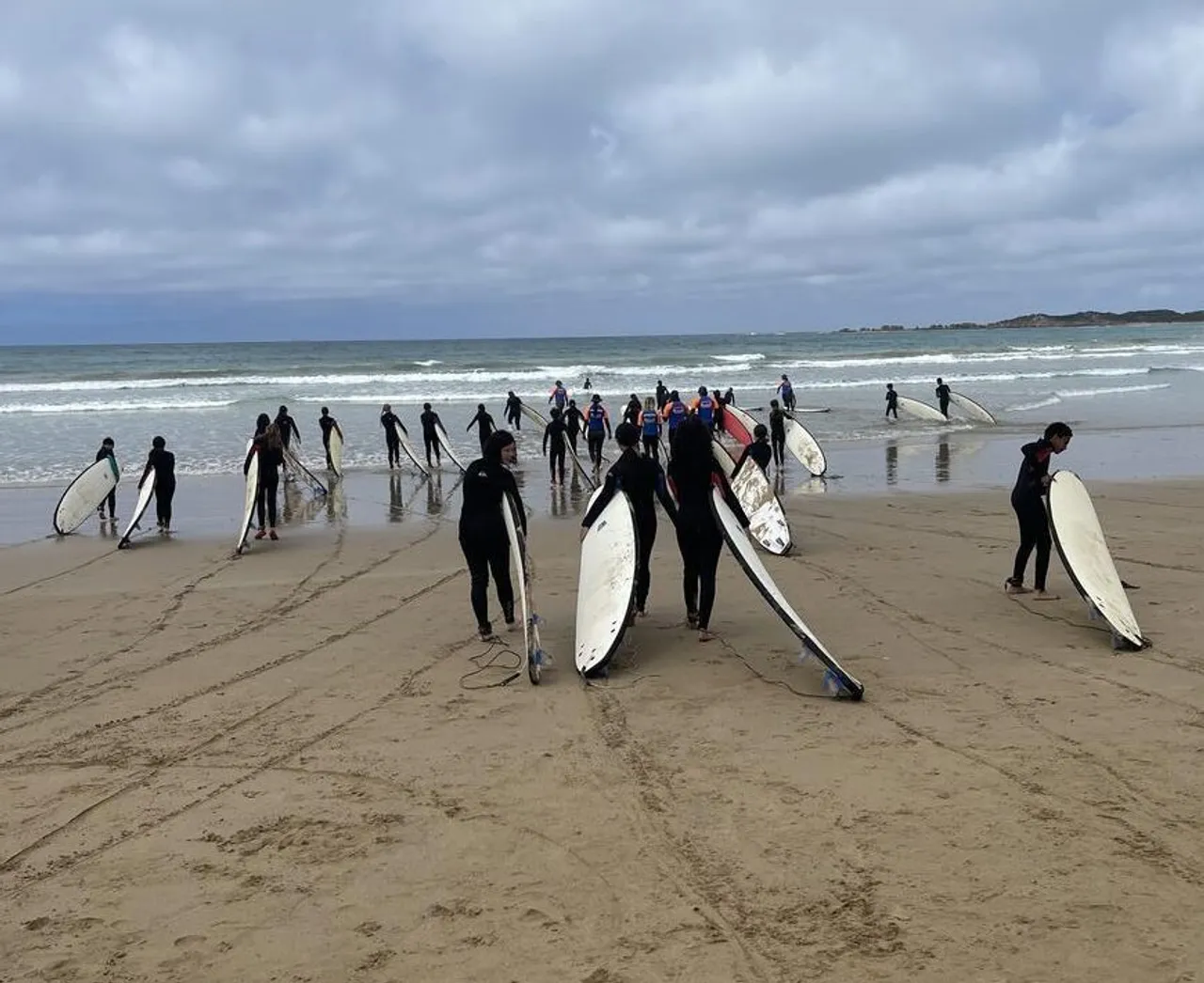 Large group of Middle School students at camp in Anglesea, all wearing wetsuits and carrying surfboards as they walk toward the ocean for a surfing lesson, ready for an outdoor adventure.