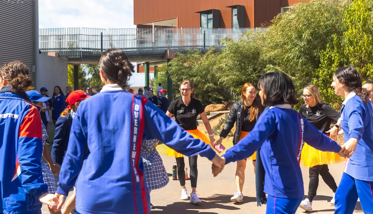 Large group of Middle School students and staff holding hands and dancing in a ring together in the outdoor courtyard at Yirramboi Campus, with the award-winning Middle School building visible in the background during Harmony Day celebrations.