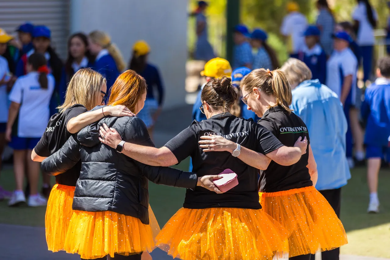 Group of teachers embracing and smiling while wearing orange tutus during Harmony Day celebrations at Overnewton, showing unity and joyful participation.