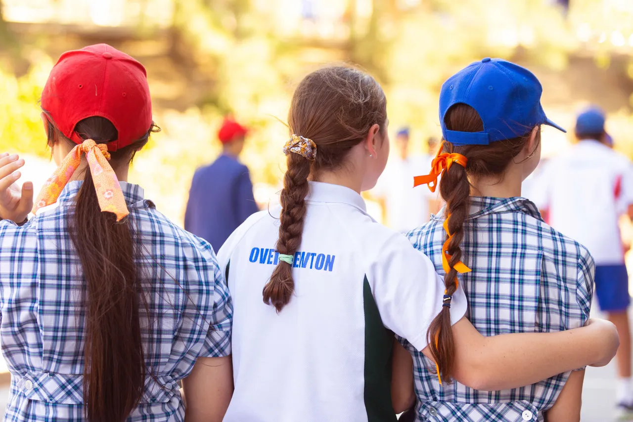 Back view of three upper primary school aged students in the Middle School at Yirramboi Campus, embracing while wearing orange hair accessories in celebration of Harmony Day.