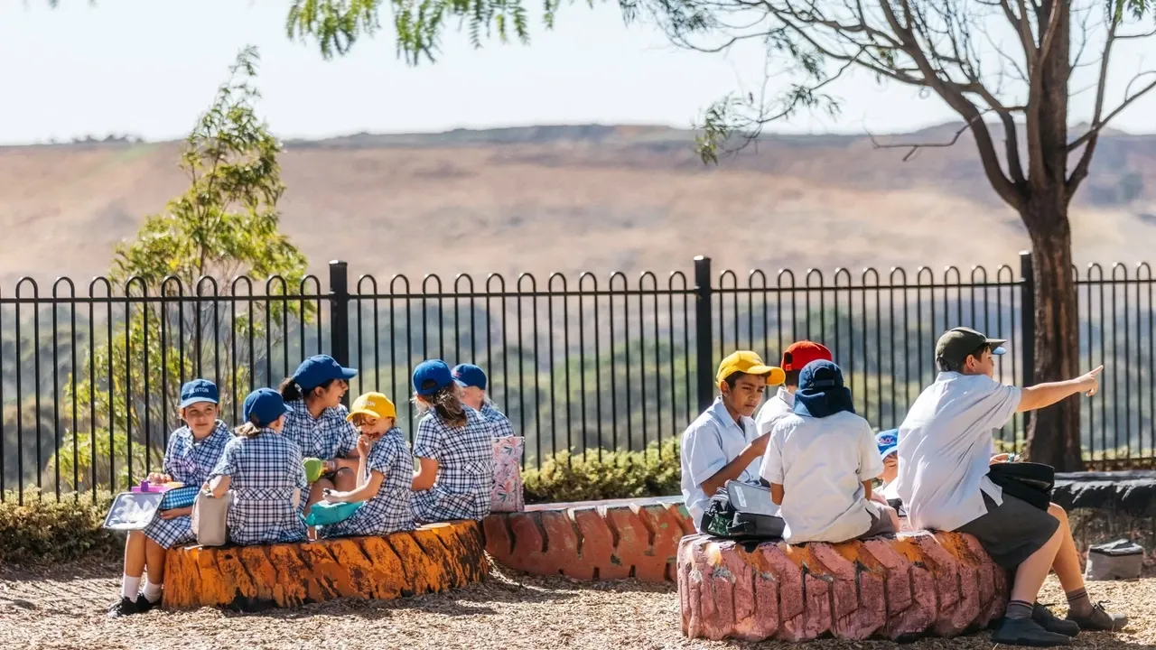 Upper primary school aged Middle School students wearing hats while eating lunch on tyres at Yirramboi Campus, with the picturesque Keilor Valley visible in the background.