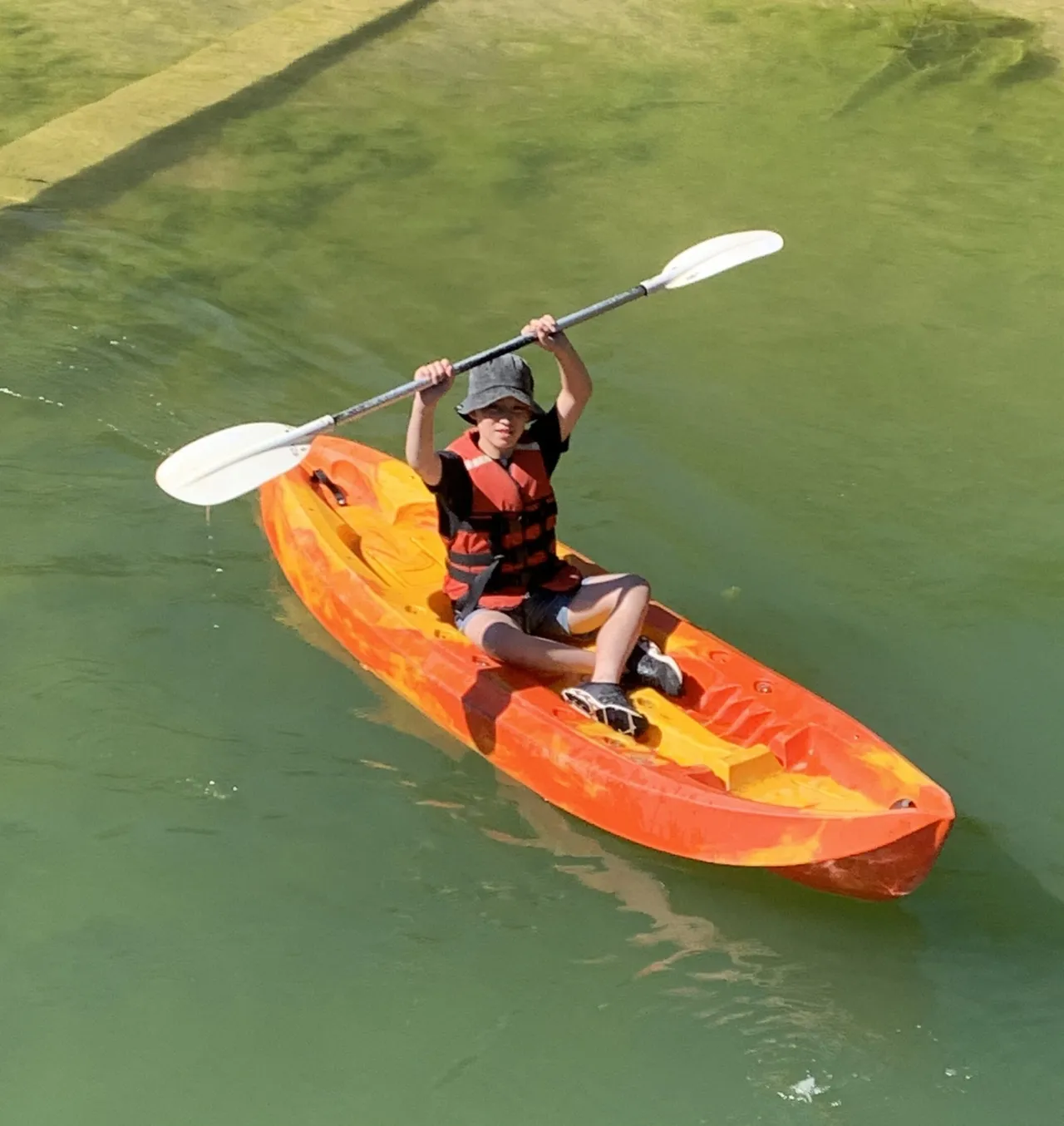 Lower secondary school aged student triumphantly holding a paddle above his head while kayaking on a lake, captured from above by a drone during the immersive Year 7 Middle School Camp program.