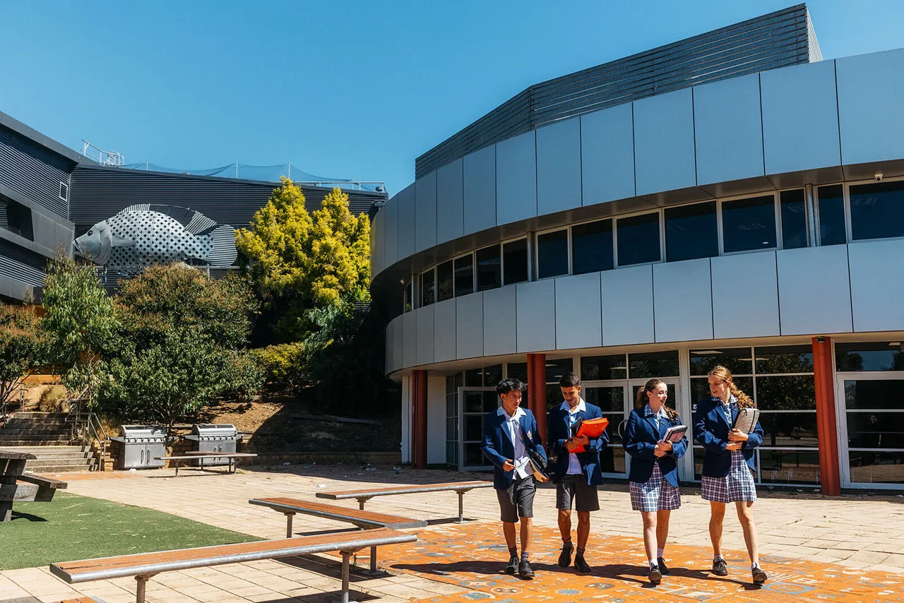 A group of four senior secondary school aged students walking with books in hand beneath a clear blue sky, framed by the sweeping architectural arc of Overnewton’s Senior School Atrium and the adjacent Sports and Performing Arts Building in the background.