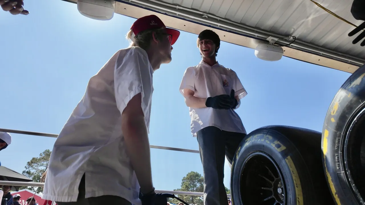 Low-angle photo of two upper secondary school aged VCE Vocational Major students smiling at each other beside a Formula 1 vehicle in bright sunlight, captured at the Driving Learning interactive educational exhibit during the Melbourne Formula 1 Grand Prix.