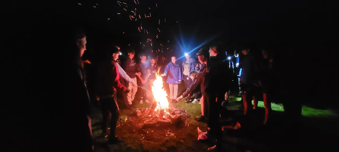 Night time photo taken during Year 9 Outdoor Education camp where middle secondary school aged students and staff faces are illuminated by the flames as they warm their hands and toast marshmallows by a roaring fire.