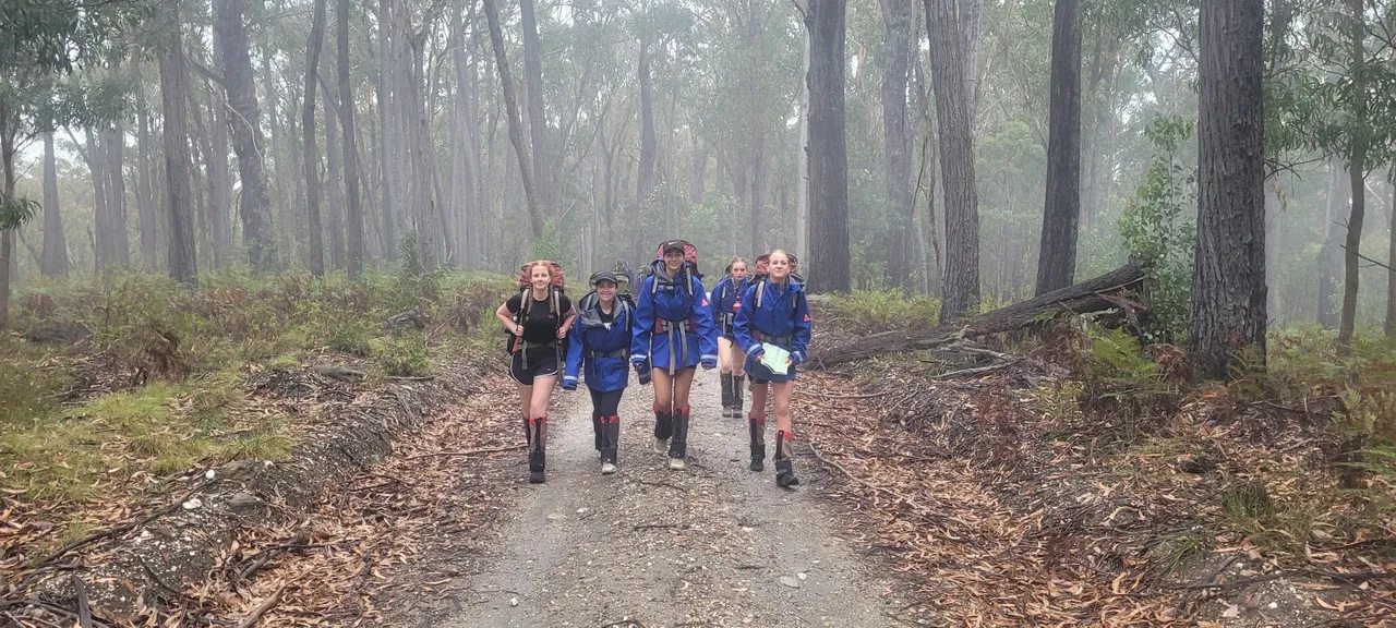 Group of middle secondary school aged students walking towards the camera on a misty forest trail, dressed in rain gear, hiking boots, and large backpacks, as they engage in the Year 9 Outdoor Education camping program.