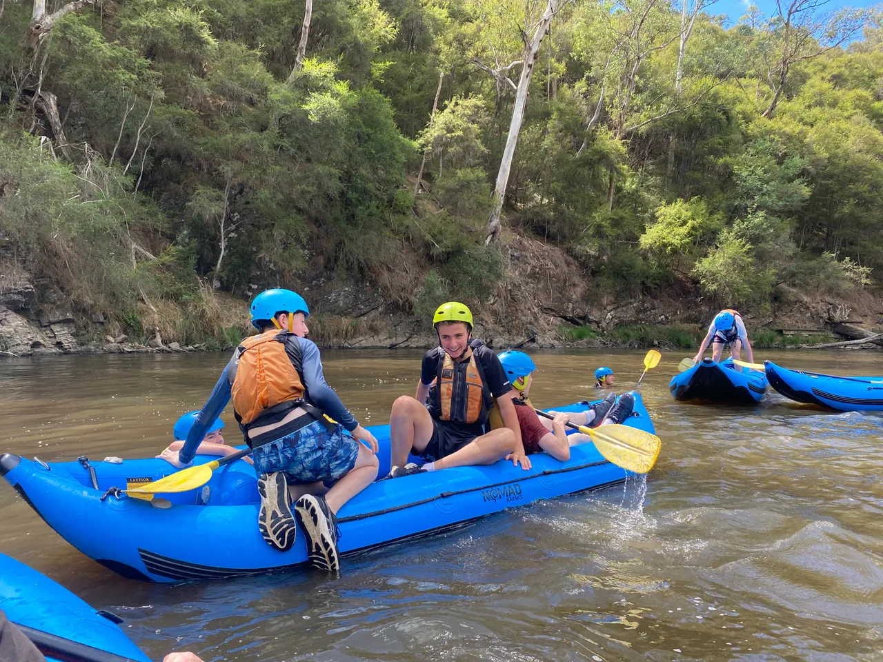 Group of middle secondary school aged students wearing helmets and wet weather gear, preparing to board a blue raft for a rafting skills session on a river in Victoria's High Country, as part of the Year 9 Outdoor Education camping program.