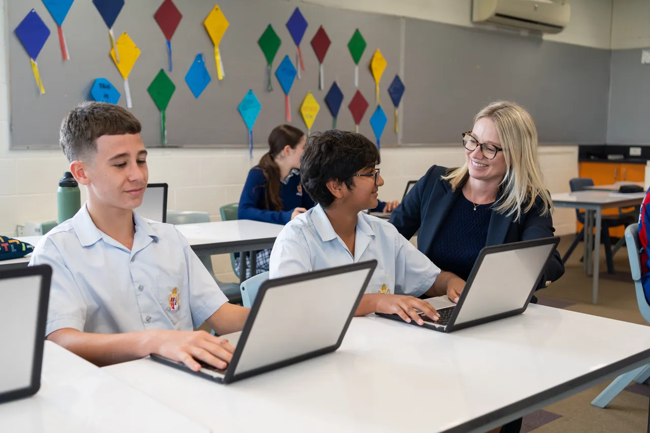 Two middle secondary school aged students engaged in learning within a bright, light-filled Year 9 Centre classroom, with their teacher crouched beside the table, offering guidance and support. A colourful display on the wall behind them adds vibrancy to the space.