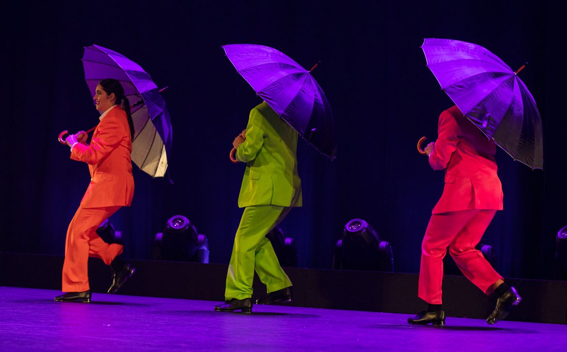 Secondary school girls perform a choreographed tap dance routine with umbrellas on stage at Penrhos College, wearing colourful suits under theatrical purple lighting during a school musical production.