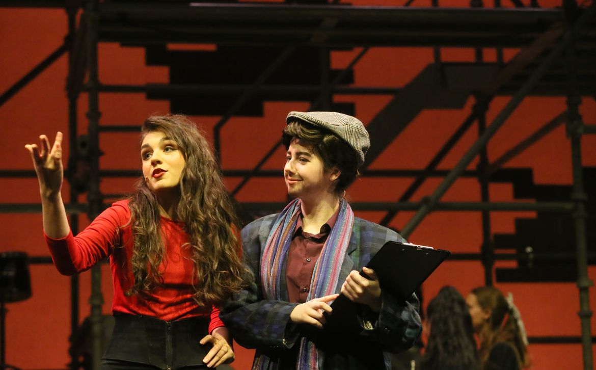 Secondary school students acting in a live stage production of the musical Fame, with two performers in costume delivering a dramatic scene against a red set and scaffolding backdrop.