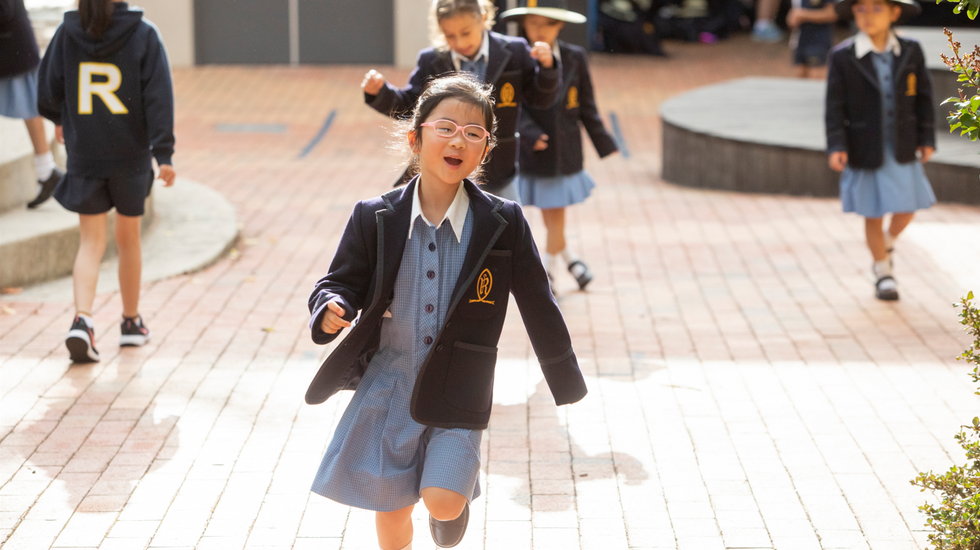 A Junior School student running and laughing in the playground at Ruyton Girls' School. She is wearing summer uniform.