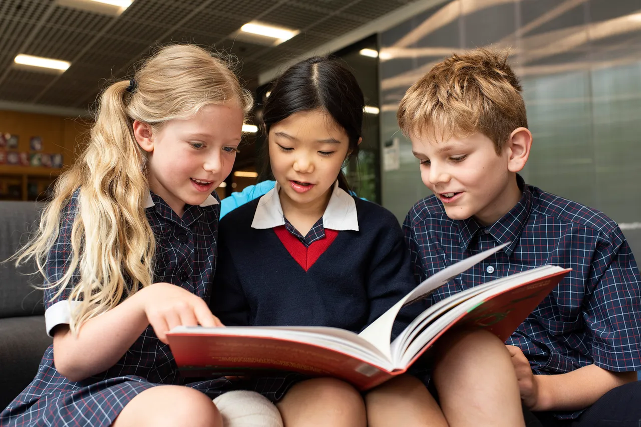 Three St Michael's Grammar School primary students huddle together reading a book in the school library.