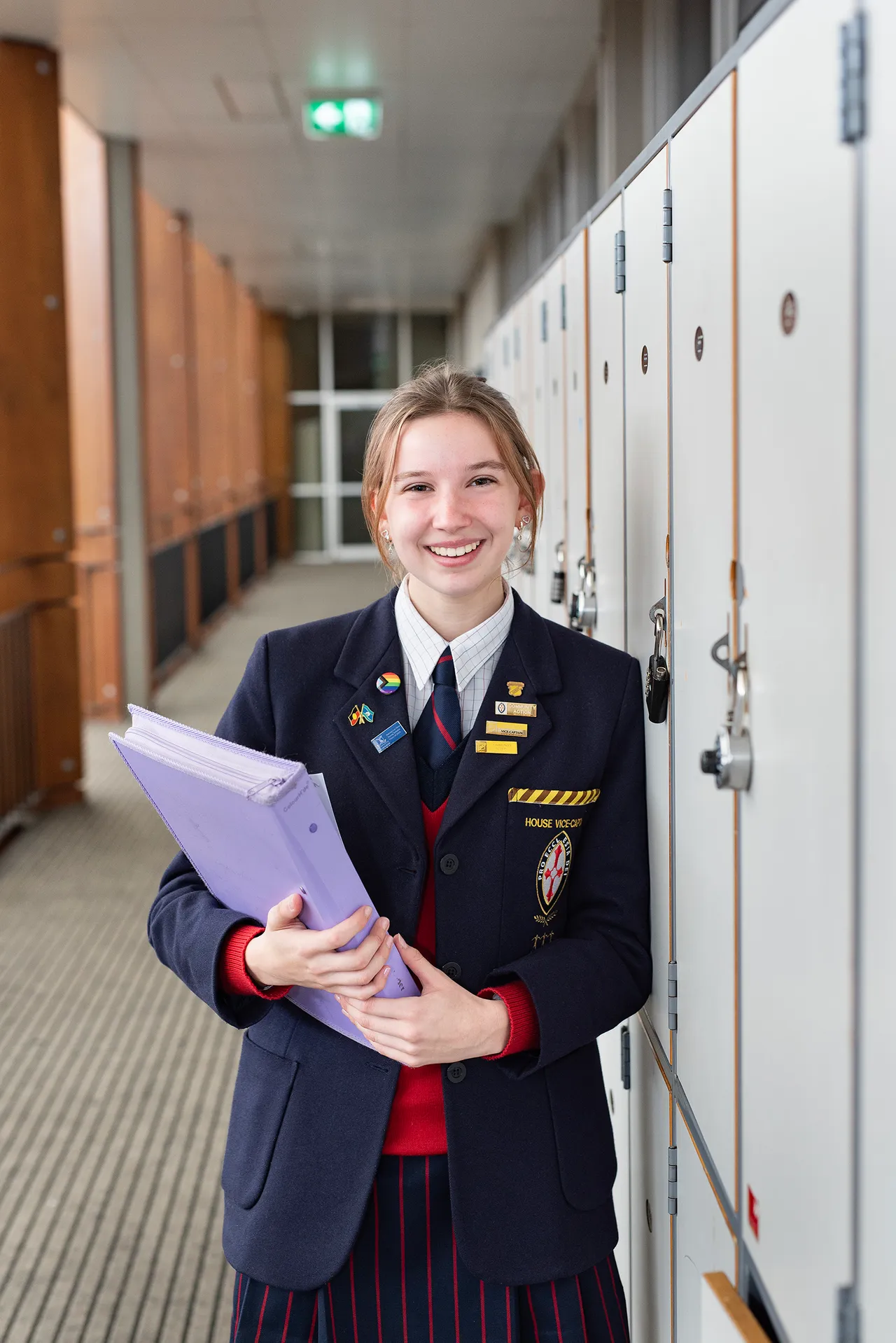 A St Michael's Grammar School senior student smiles at the camera while standing at her locker in a school corridor, holding a folder.