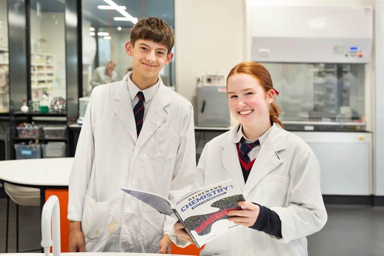 Two senior St Michael's Grammar School students in white lab coats smile at the camera in a science laboratory, one holding a chemistry textbook.