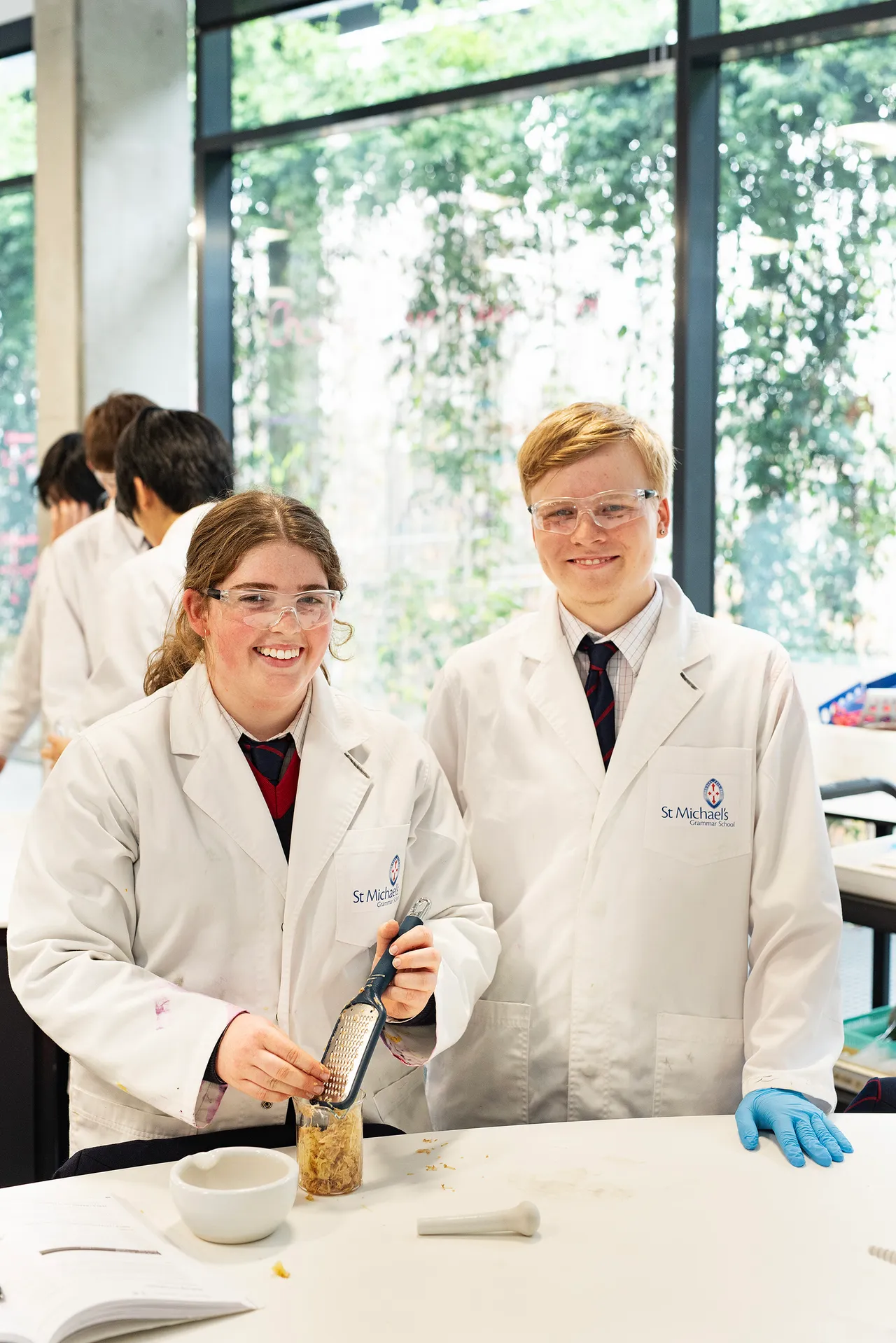 Two St Michael's Grammar School senior students in white lab coats and safety glasses smile in the science facilities.