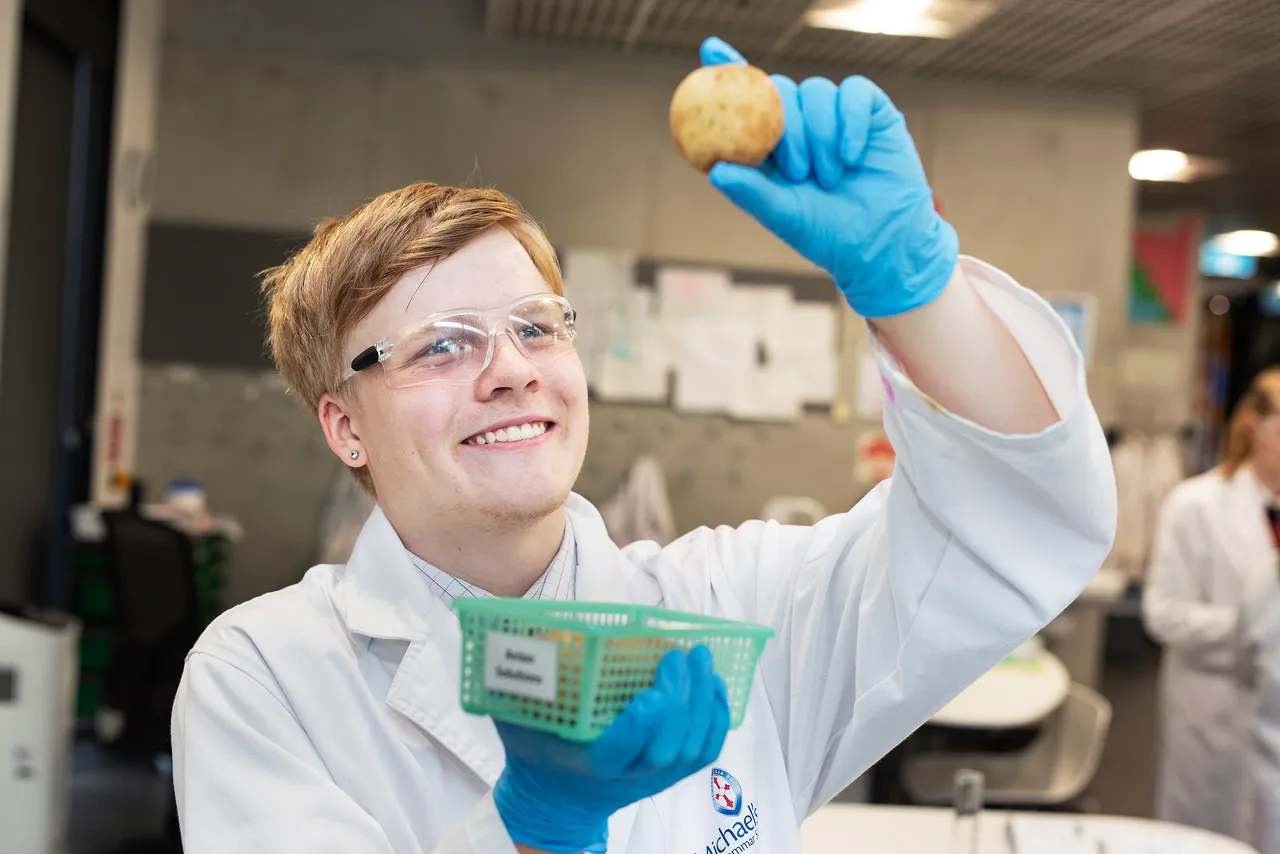 A St Michael's Grammar School student in a white lab coat and safety glasses holds up a specimen in a science laboratory.