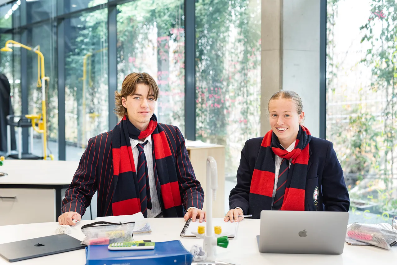 Two St Michael's Grammar School senior students wearing school scarves smile at the camera while working at a desk in a light-filled learning space.