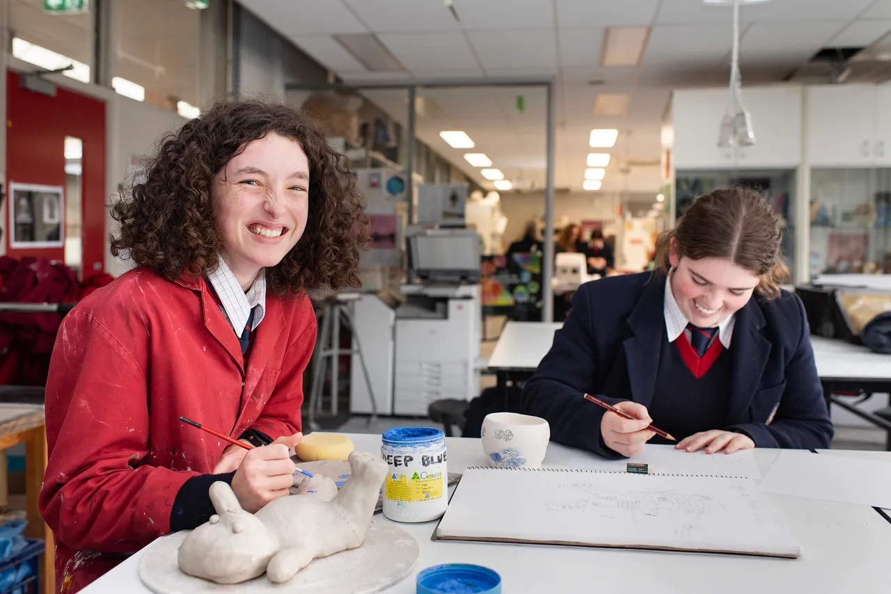 Two St Michael's Grammar School senior students work at a desk in a design classroom, one smiling at the camera while the other focuses on their work.