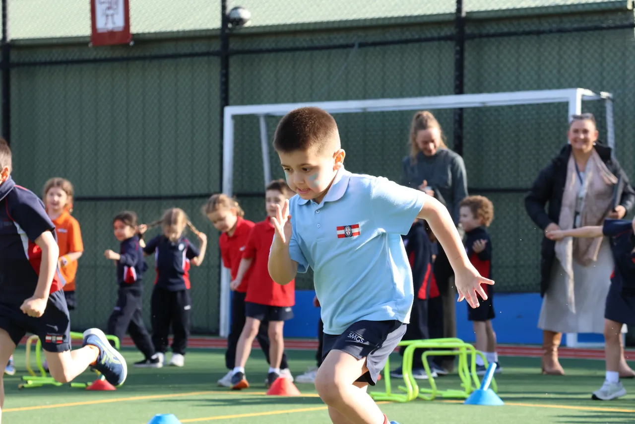 A young St Michael's Grammar School student sprints during an outdoor PE class on the school's sports court, with classmates and teachers active in the background.