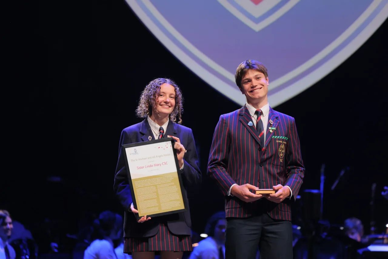 Two St Michael's Grammar School senior students stand on stage holding awards during a formal school presentation ceremony.