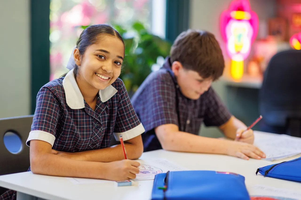 A St Michael's Grammar School student smiles at the camera while working at her desk, with a classmate writing beside her in a bright, decorated classroom.