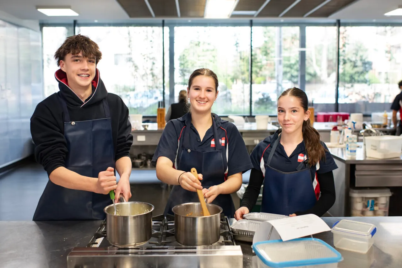 Three St Michael's Grammar School students in aprons smile at the camera while cooking together in the school's food technology kitchen.