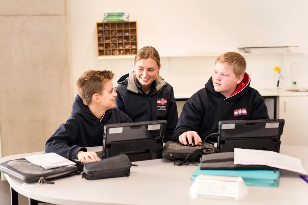 A St Michael's Grammar School teacher smiles with two students as they work together on laptops during a classroom lesson.