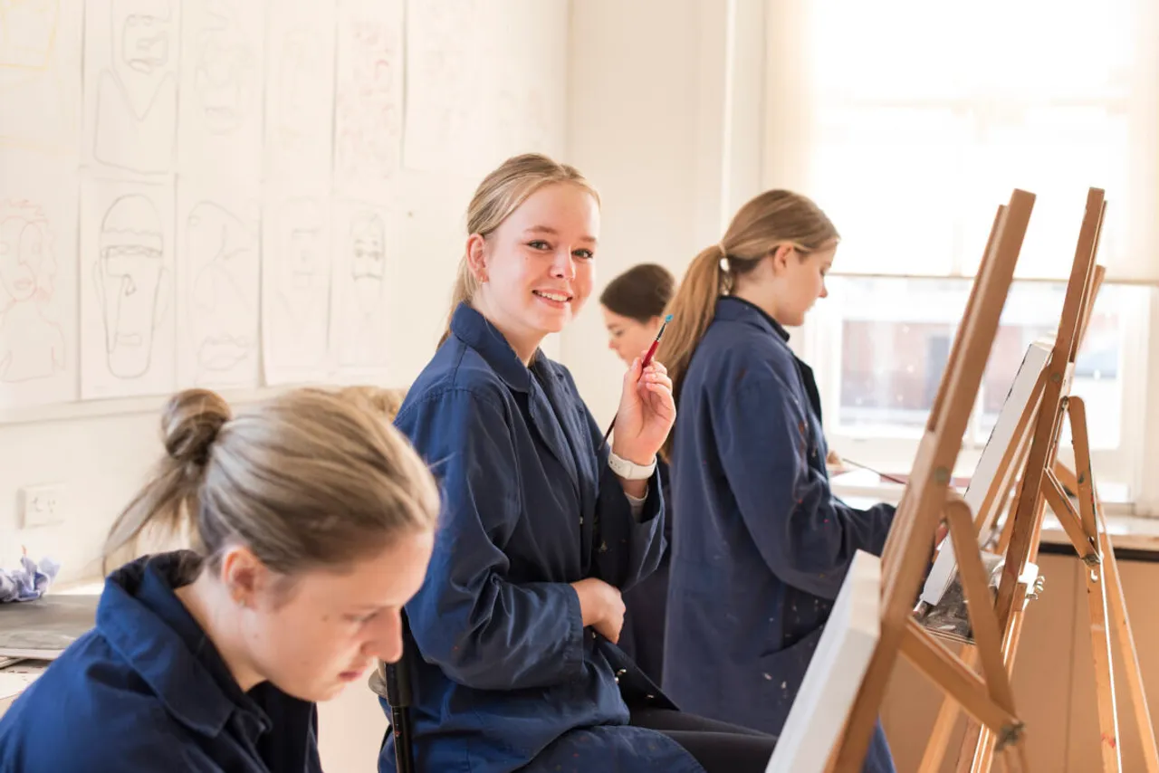 St Michael's Grammar School students in art smocks work at easels during a studio art class, one pausing to smile at the camera.
