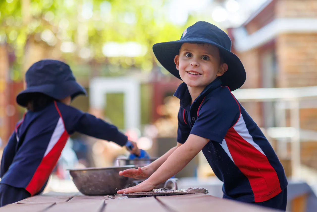 A young St Michael's Grammar School student in a sun hat smiles while playing outdoors at a water and mud sensory table.