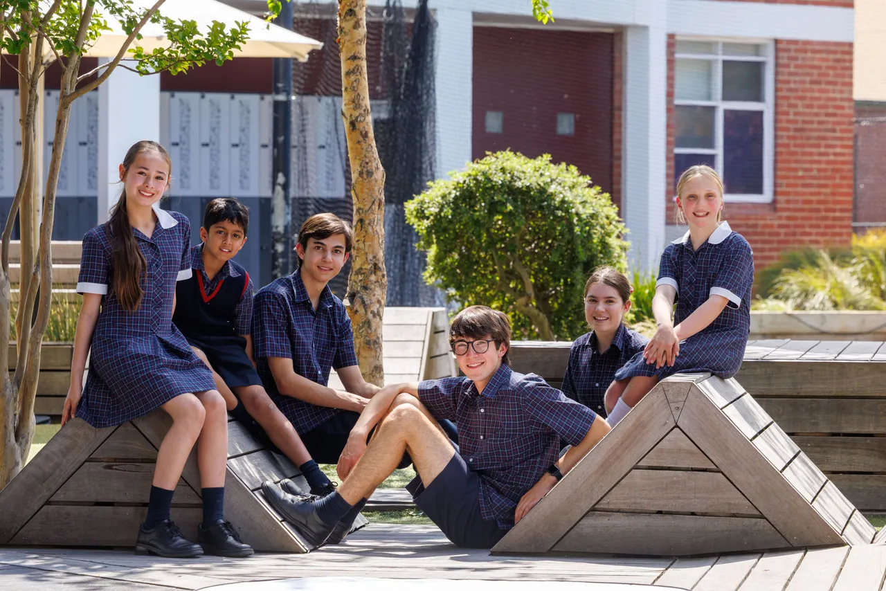A group of St Michael's Grammar School students in uniform relax together on outdoor seating in the school's courtyard, smiling at the camera.