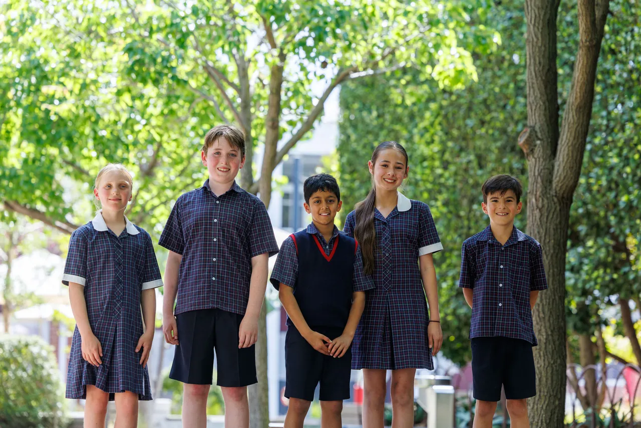 Five St Michael's Grammar School primary students in summer uniform stand together outdoors on the school grounds, smiling at the camera.