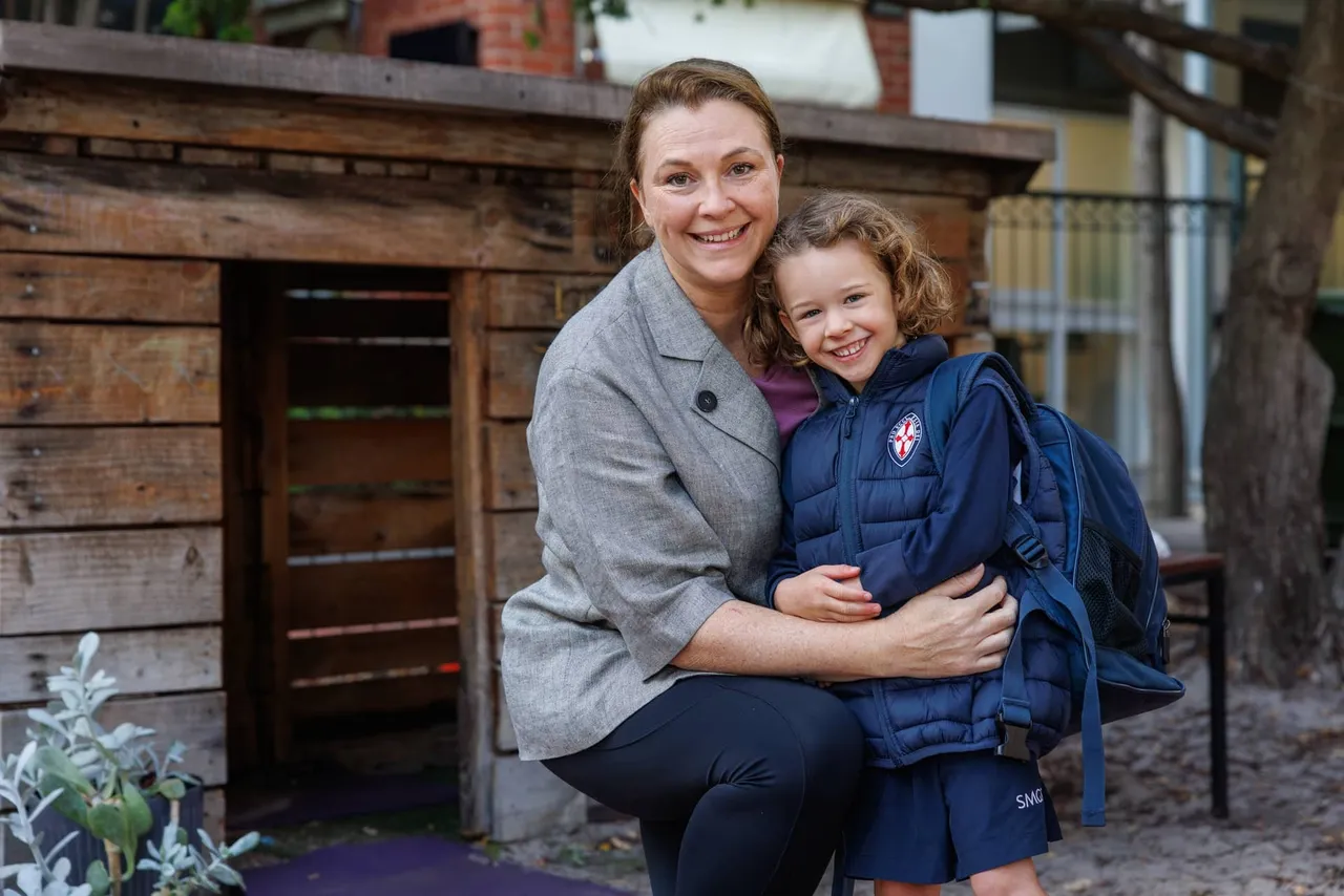 The image shows a mother and young child (daughter) in school uniform, smiling together outdoors in what appears to be a natural timber playground setting.