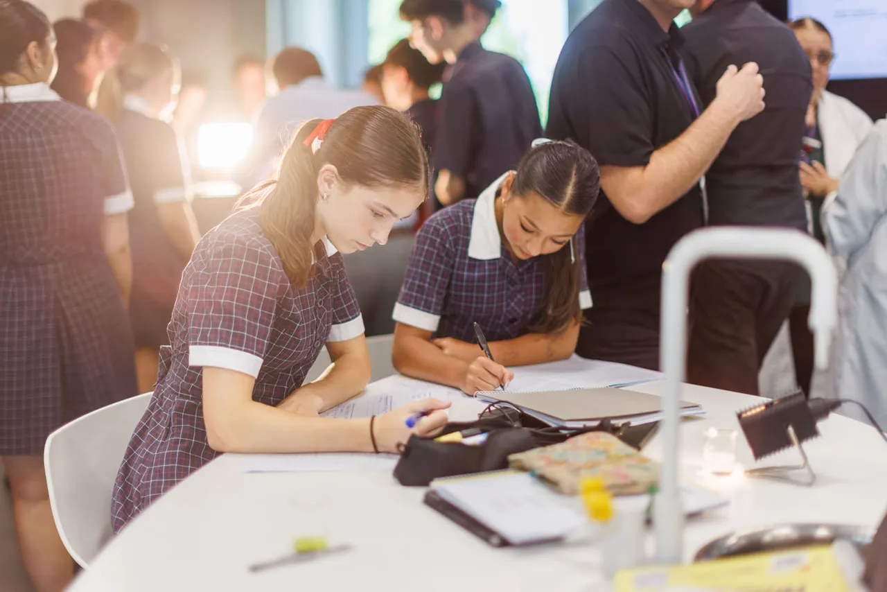 Two St Michael's Grammar School students in uniform work together at a desk during a science class, with one student writing notes while the other observes.
