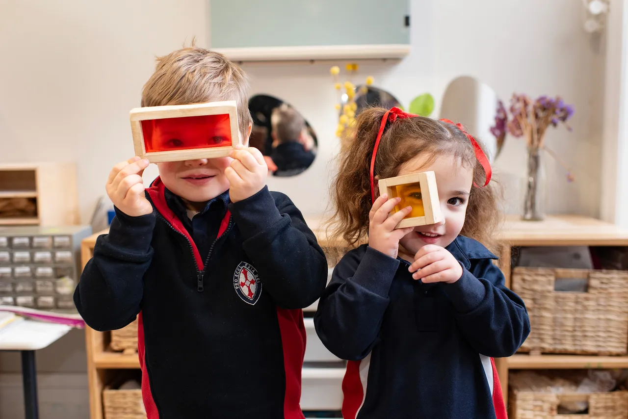 Two young St Michael's Grammar School students hold wooden frame viewers up to their eyes during an ELC activity, both smiling as they explore.