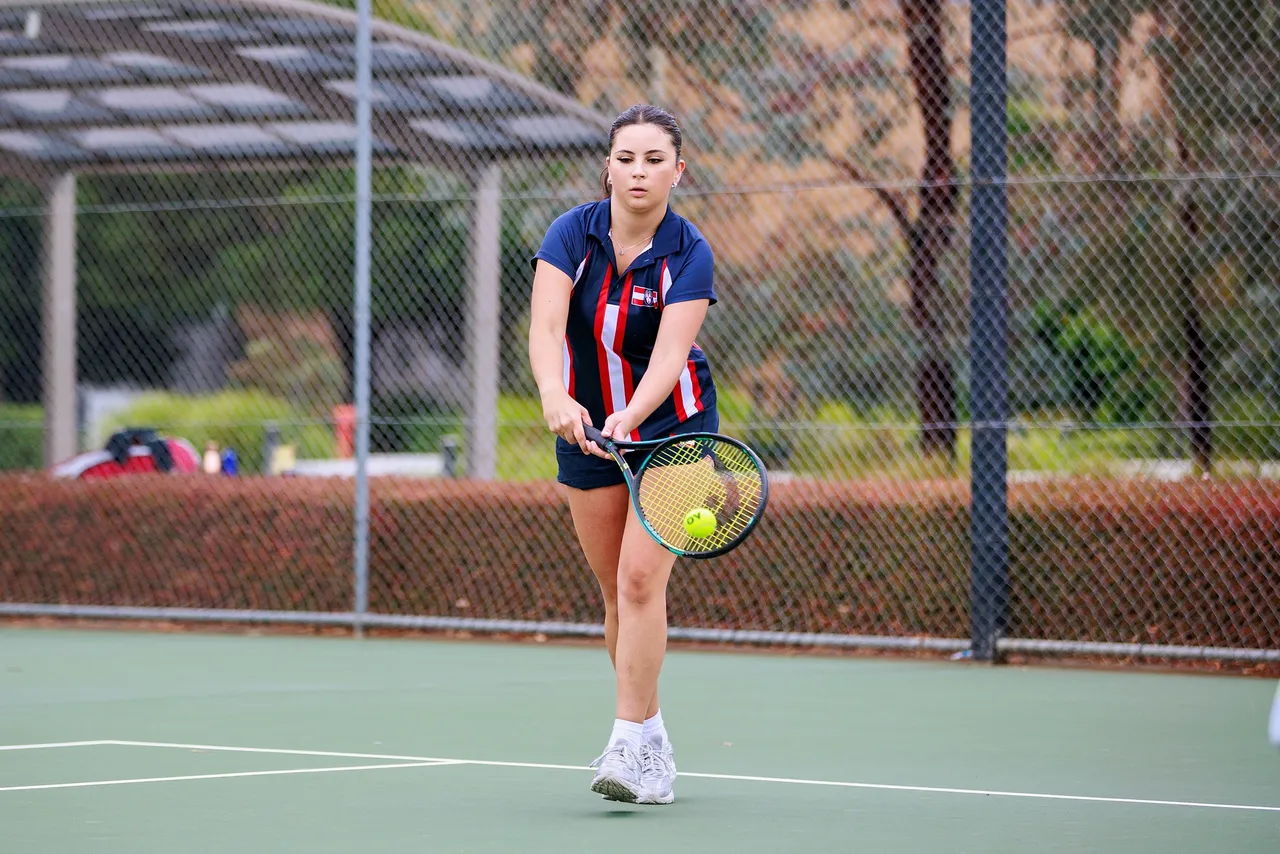A St Michael's Grammar School student in sport uniform plays tennis on an outdoor court during a school sports match.