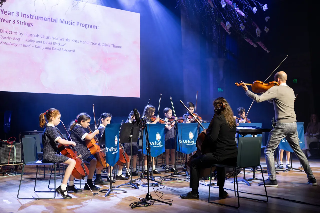 A conductor leads St Michael's Grammar School student string musicians on stage during a formal instrumental music concert.