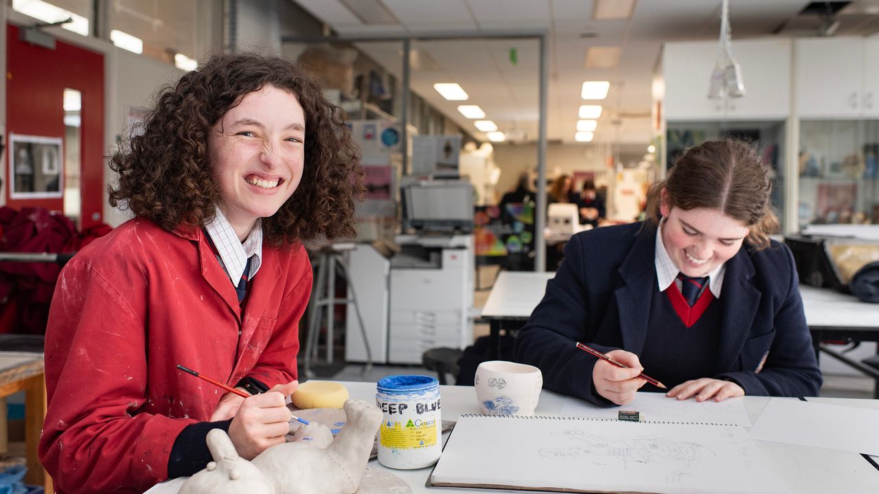 Two St Michael's Grammar School senior students work at a desk in a design classroom, one smiling at the camera while the other focuses on their work.