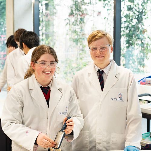 Two St Michael's Grammar School senior students in white lab coats and safety glasses smile in the science facilities.
