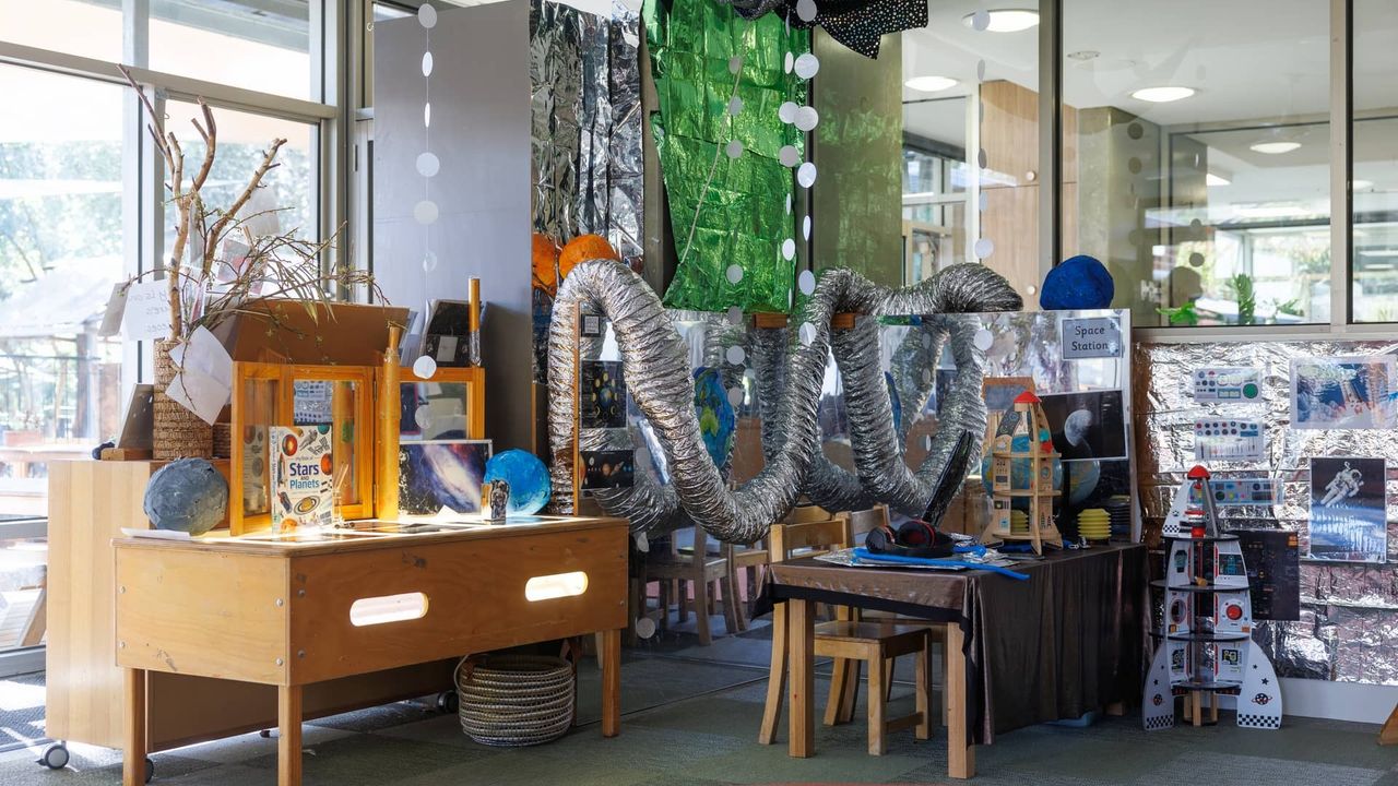 A richly decorated space-themed learning environment in the St Michael's Grammar School ELC, featuring a light table, planet models, a cardboard rocket and imaginative loose materials for exploration.