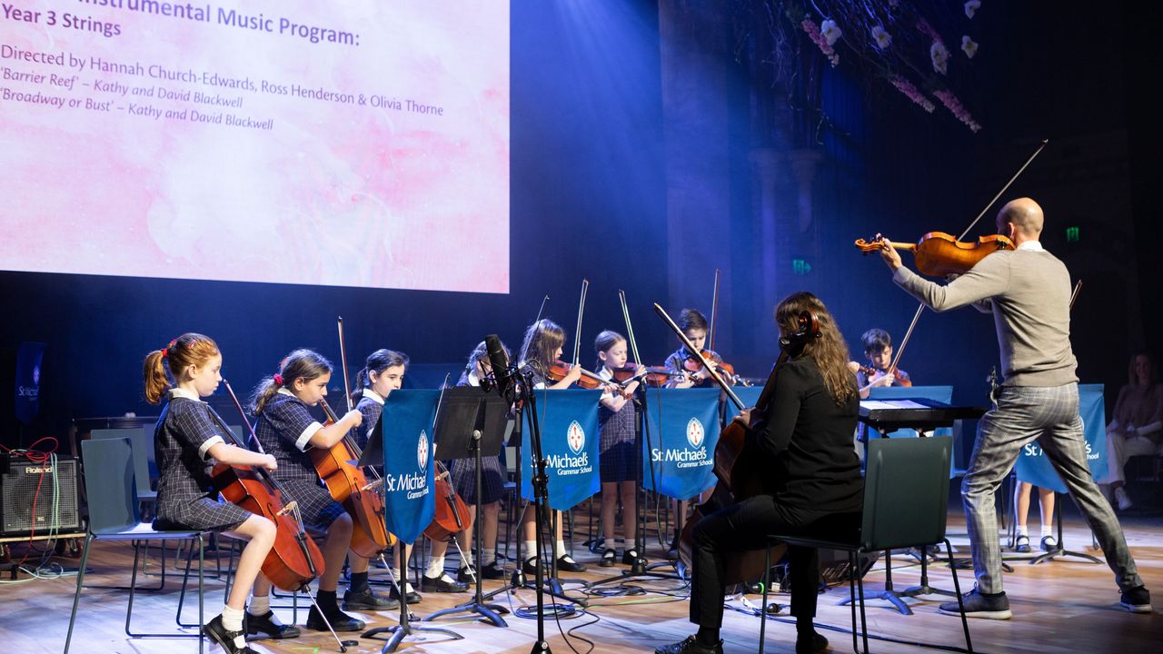 A conductor leads St Michael's Grammar School student string musicians on stage during a formal instrumental music concert.